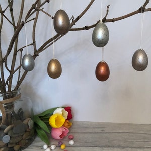 May include: A branch with six hanging Easter eggs in different metallic colours: gold, silver, copper, and grey. The eggs are hanging from white string. There are also a few small, speckled Easter eggs on a wooden surface.