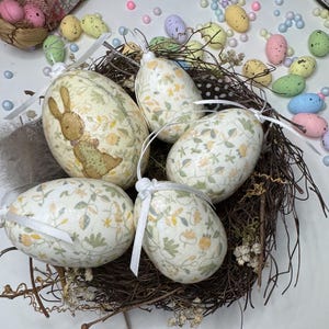 May include: Five decorative Easter eggs in a bird's nest. The eggs are white with a floral pattern and are tied with white ribbon.