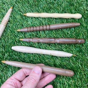 May include: A selection of handcrafted wooden pens in various styles and wood tones, including light and dark brown, and natural wood colours. The pens have gold-coloured metal tips, displayed on green grass.