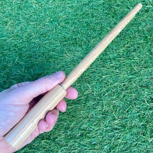 May include: A light-coloured wooden wand with a cylindrical handle and a tapered shaft. The wand is held against a background of green grass. The wand is made of light-coloured wood and has a smooth finish.