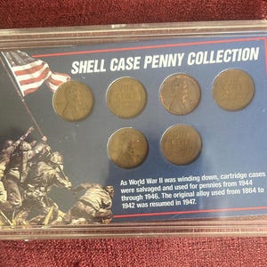 Puede incluir: Una caja de plástico transparente que contiene una colección de monedas Shell Case. La exhibición presenta seis centavos de cobre y un fondo azul con una imagen de soldados izando la bandera estadounidense. El texto dice "SHELL CASE PENNY COLLECTION."