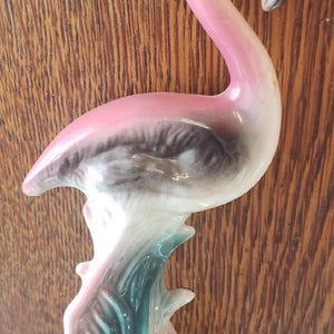 Puede incluir: Figurita de flamenco de cerámica en tonos rosa, negro, blanco y verde azulado. El flamenco se alza sobre una base en forma de árbol, con un cuello largo y curvado y un pico negro. Es un objeto decorativo.