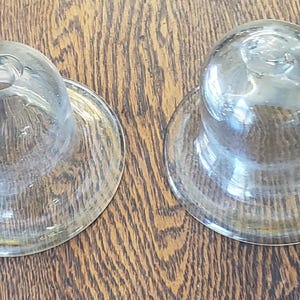 May include: Two clear glass domes with wide, flat circular bases. Each piece features a smooth, rounded top section that transitions into a flared rim. The glass is transparent and reflects light, showing a simple, functional, and symmetrical design.