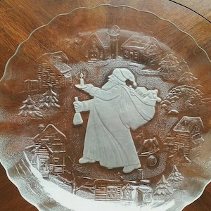 Könnte beinhalten: Ein klarer Weihnachtsteller aus Glas mit einem erhabenen Reliefdesign. Der Teller zeigt eine Darstellung des Weihnachtsmanns, der eine Kerze und eine Tasche trägt und durch eine Winterdorflandschaft mit Häusern und Bäumen geht. Der Teller hat einen gewellten Rand.