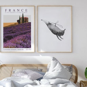 May include: Two framed prints hanging on a white wall above a bed with white linens and a wicker headboard. The print on the left features a lavender field in France with the text "FRANCE C'est toujours le bon moment. LAVENDER SESSION, ARDECHE JULY 2021 CHLOE TIMMER". The print on the right features a black and white illustration of a bird with a propeller on its head.