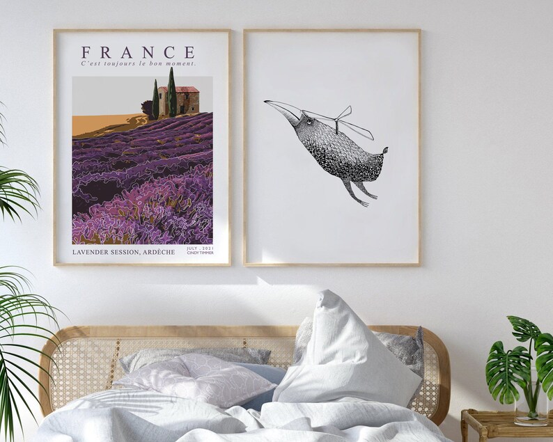 May include: Two framed prints hanging on a white wall above a bed with white linens and a wicker headboard. The print on the left features a lavender field in France with the text "FRANCE C'est toujours le bon moment. LAVENDER SESSION, ARDECHE JULY 2021 CHLOE TIMMER". The print on the right features a black and white illustration of a bird with a propeller on its head.