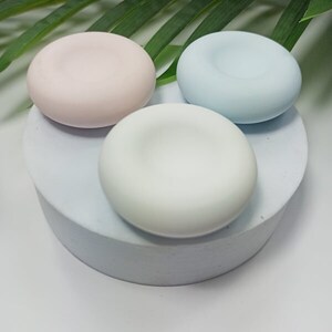 May include: Three pastel-coloured, round soap dishes. The dishes are light pink, light blue, and white, each with a shallow, circular indentation. They rest on a light grey, cylindrical pedestal, with a green leaf in the background.