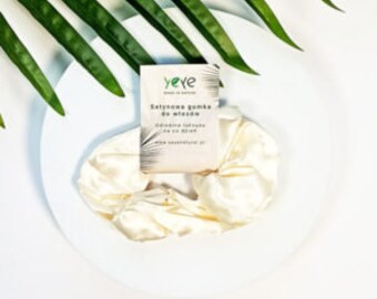 May include: A white scrunchie and a small rectangular card with text are displayed on a white plate, with green leaves in the background. The card has the text "yeye" and "Satynowa gumka do włosów".