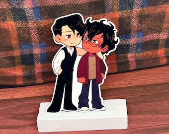 May include: A white wooden stand with two cartoon figures. The figure on the left is wearing a black suit and a white shirt with a black vest. The figure on the right is wearing a red jacket, blue jeans, and green glasses.