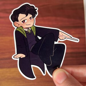 May include: A cartoon illustration of a young person with black hair wearing a black and green robe, sitting with one leg crossed over the other, holding a wand in their right hand.