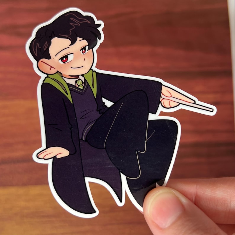 May include: A cartoon illustration of a young person with black hair wearing a black and green robe, sitting with one leg crossed over the other, holding a wand in their right hand.