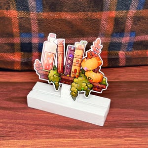 Puede incluir: Un soporte de madera blanco con una ilustración colorida de un estante con libros, una botella y calabazas. El estante está decorado con enredaderas verdes y purpurina.