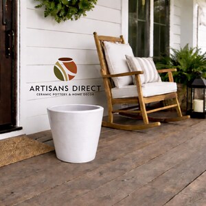 May include: A white ceramic planter sits on a wooden porch. A wooden rocking chair with white cushions and a striped pillow is nearby. The porch has a dark brown door and a welcome mat. The image includes the text "ARTISANS DIRECT CERAMIC POTTERY & HOME DECOR."