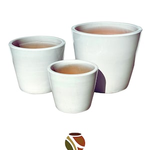 May include: Three white ceramic planters of varying sizes. The planters have a glossy finish and a simple, modern design. The interior of each planter is a natural terracotta color. The logo "ARTISANS DIRECT CERAMIC POTTERY & HOME DECOR" is at the bottom.