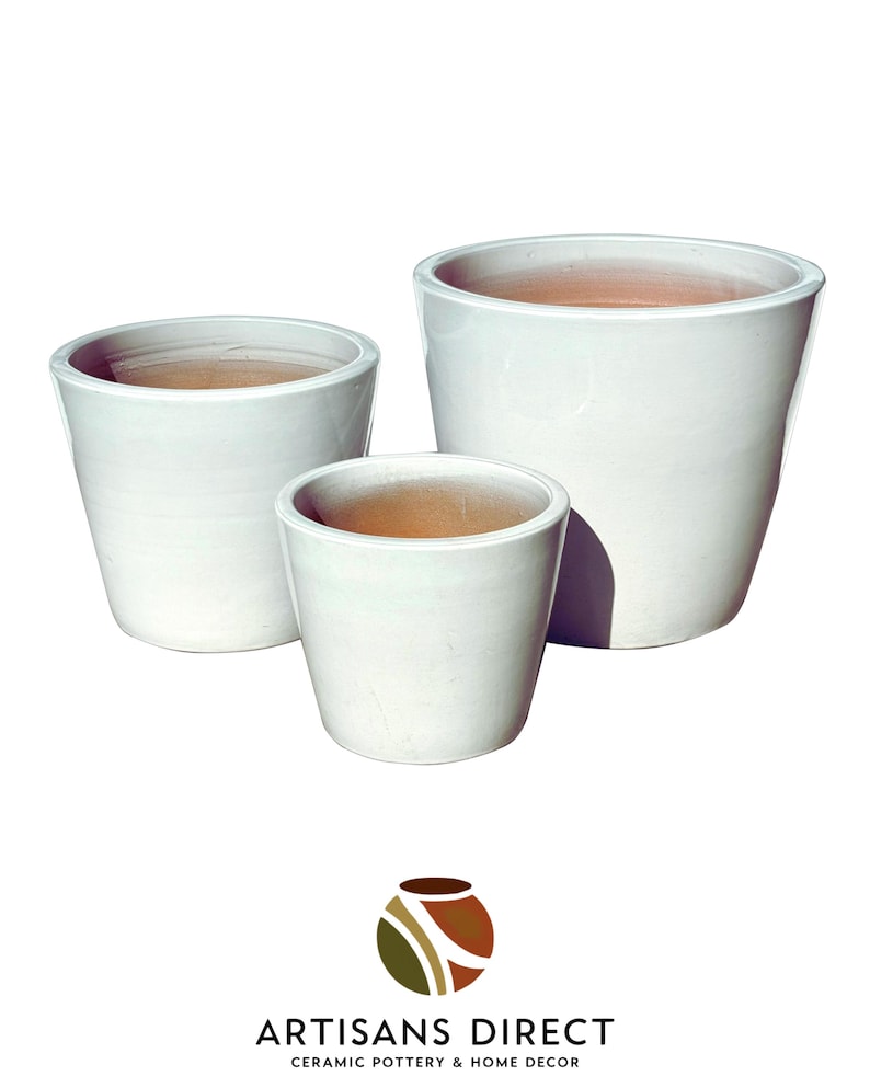 May include: Three white ceramic planters of varying sizes. The planters have a glossy finish and a simple, modern design. The interior of each planter is a natural terracotta color. The logo "ARTISANS DIRECT CERAMIC POTTERY & HOME DECOR" is at the bottom.