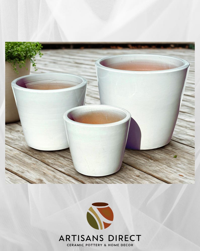 May include: Three white ceramic planters of varying sizes are arranged on a wooden surface. The planters have a glossy finish and a simple, modern design. The interior of each planter is a natural terracotta color. The logo "ARTISANS DIRECT CERAMIC POTTERY & HOME DECOR" is at the bottom.