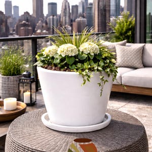 May include: A large white ceramic planter filled with green plants and white flowers, set on a woven table. The background features a cityscape with tall buildings and a clear sky. The planter is on a white saucer.