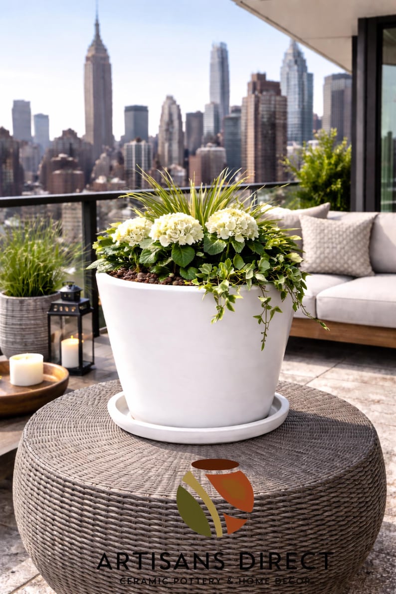 May include: A large white ceramic planter filled with green plants and white flowers, set on a woven table. The background features a cityscape with tall buildings and a clear sky. The planter is on a white saucer.