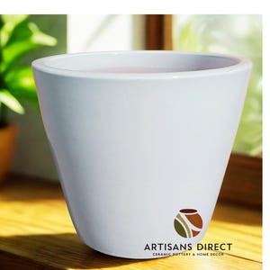 May include: A white ceramic planter with a slightly flared shape. The planter has a smooth, glossy finish and a wide opening. The logo "ARTISANS DIRECT CERAMIC POTTERY & HOME DECOR" is printed on the front. The planter is suitable for indoor or outdoor use.