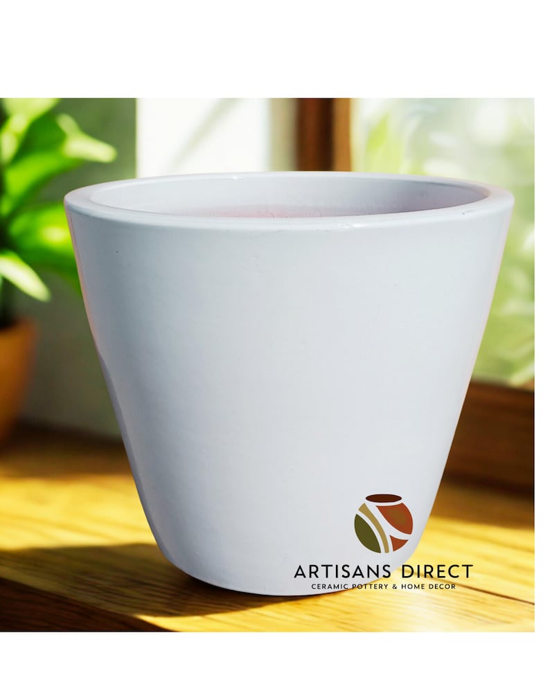 May include: A white ceramic planter with a slightly flared shape. The planter has a smooth, glossy finish and a wide opening. The logo "ARTISANS DIRECT CERAMIC POTTERY & HOME DECOR" is printed on the front. The planter is suitable for indoor or outdoor use.