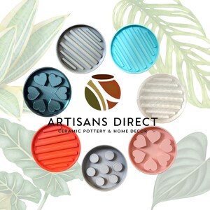 May include: Assortment of seven colorful ceramic coasters with various designs, including stripes, hearts, and circles. The coasters are arranged in a circle around the logo "ARTISANS DIRECT CERAMIC POTTERY & HOME DECOR".