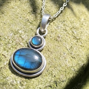 Puede incluir: Collar con colgante de plata con una piedra grande ovalada y una pequeña redonda de labradorita azul. Las piedras están engastadas en un bisel de plata y conectadas por una cadena de plata. El colgante se muestra sobre una superficie verde texturizada.