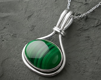 May include: A silver pendant necklace featuring an oval-shaped malachite gemstone with deep green banded patterns. The stone is set in a polished silver wire frame that loops at the top to connect to a delicate silver chain link necklace.