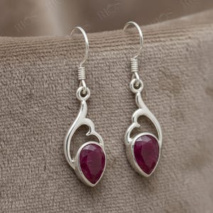 May include: A pair of silver drop earrings featuring a teardrop-shaped deep red gemstone set in a polished metal frame with a curved openwork design. The earrings hang from silver fishhook wires, showcasing a faceted surface on the stones.