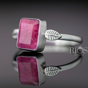 May include: A silver ring featuring a square, red ruby gemstone and leaf accents. The ring is displayed on a reflective surface.