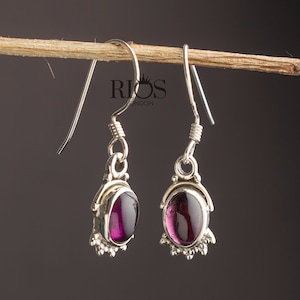 Könnte beinhalten: Ein Paar silberne Ohrhänger mit violetten, ovalen Edelsteinen. Die Ohrringe haben ein filigranes Design mit kleinen Details um die Edelsteine herum.