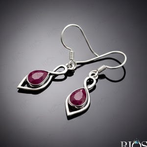 May include: A pair of silver drop earrings featuring faceted, teardrop-shaped deep red gemstones set within a polished silver frame. The design includes a delicate infinity-style loop connecting the gemstone setting to the silver ear wire hooks.
