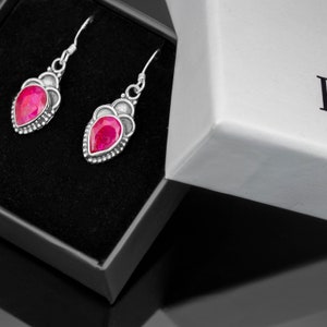 May include: A pair of silver earrings with pink gemstone drops, displayed in a black and white gift box.