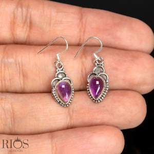 Könnte beinhalten: Ein Paar silberne Ohrhänger mit violetten Amethyst-Edelsteinen. Die Ohrringe haben eine Tropfenform und sind mit einem silbernen Filigranmuster verziert.