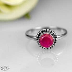 Pode incluir: Um anel de prata com uma vibrante pedra preciosa de rubi redonda. O rubi está definido em uma moldura com contas. O anel é exibido em uma superfície reflexiva. A marca "RIOS LONDON" é visível.