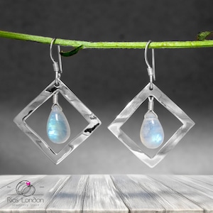 May include: Silver earrings with a square frame and a teardrop-shaped moonstone pendant. The earrings are hanging from a green branch.