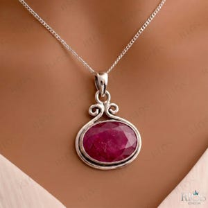 Puede incluir: Un collar de plata con una gran piedra preciosa de rubí de color rosa intenso, de forma ovalada y facetada, engastada en un marco de plata pulida con volutas decorativas en la parte superior. El colgante cuelga de una delicada cadena de plata.