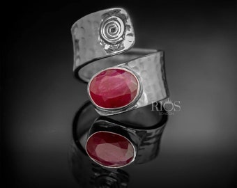 May include: A silver ring with a hammered finish and a red oval gemstone. The ring has a spiral design on one side and the text "RIOS LONDON" on the other side.