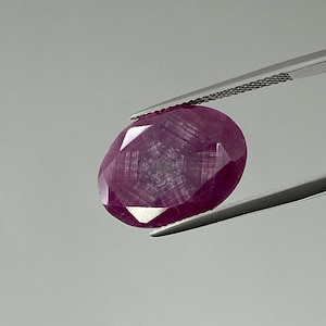 Puede incluir: Una piedra preciosa ovalada de color rojo oscuro con un corte facetado. La piedra preciosa está sujeta por unas pinzas.