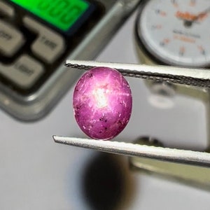 Op de afbeelding: Een close-up van een roze robijn edelsteen met een stervormige insluiting. De robijn wordt vastgehouden in een pincet en wordt gemeten met een meetinstrument.