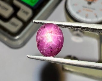 May include: A close-up of a pink ruby gemstone with a star-shaped inclusion. The ruby is held in tweezers and is being measured with a gauge.