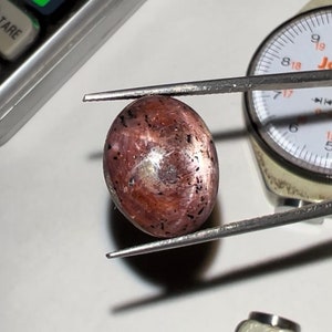 Puede incluir: Una gema ovalada pulida, de color marr&oacute;n rojizo con inclusiones oscuras. Se sujeta con pinzas, con un indicador de esfera y una calculadora en el fondo. La superficie de la piedra refleja la luz, mostrando su brillo.