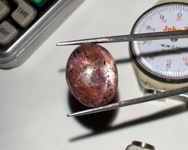 Puede incluir: Una gema ovalada pulida, de color marr&oacute;n rojizo con inclusiones oscuras. Se sujeta con pinzas, con un indicador de esfera y una calculadora en el fondo. La superficie de la piedra refleja la luz, mostrando su brillo.