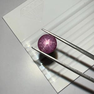 May include: A round, purplish-pink gemstone, held by silver tweezers, displays a star-like effect. The gem sits on a white, ridged surface with a diamond-shaped logo. The lighting highlights the gem's facets and the tweezers' metallic sheen.