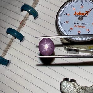 Puede incluir: Una gema redonda de color rosa púrpura sujeta con pinzas, junto a un calibrador de gemas. El calibrador de gemas tiene una esfera blanca con números negros y el texto "Johar Gem Gauge". Varios clips azules son visibles en el fondo.