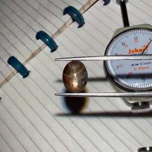 May include: A brown, oval gemstone is held by a dial caliper, displaying measurements. The gemstone has a speckled, reflective surface. The caliper's face shows measurements in millimetres. A wooden stick with blue clips is in the background.