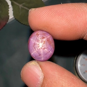 May include: A round, cabochon-cut star ruby gemstone with a distinct six-rayed asterism pattern visible on its translucent, purplish-pink surface. The stone displays natural internal inclusions and a smooth, polished dome shape.