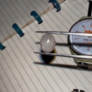 May include: A light pink, oval-shaped gemstone is held by a caliper, with a dial gauge in the background. The gemstone is being measured. The background includes a lined notebook.