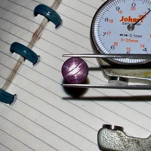 May include: A purplish-pink gemstone, spherical in shape, is held by a gem gauge. The gauge has a white dial with black numbers and the text "Johan Gem Gauge". The gemstone is resting on lined paper. The gauge measures in millimeters.