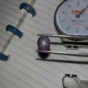 以下が含まれることがあります： ピンセットで保持された紫がかったピンク色のスターサファイアの宝石、Johar Gem Gaugeの隣。 宝石には六条の星型模様があります。 ゲージはミリメートルで測定します。