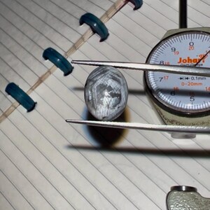May include: A close-up of a gemstone being measured with a dial gauge. The gemstone is a light gray color with a patterned surface. The gauge reads in millimeters and has the brand name "Joha" visible.